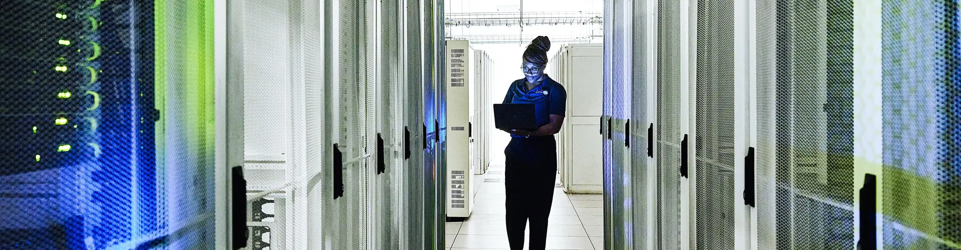 woman is walking down a hallway in a server room looking at her laptop