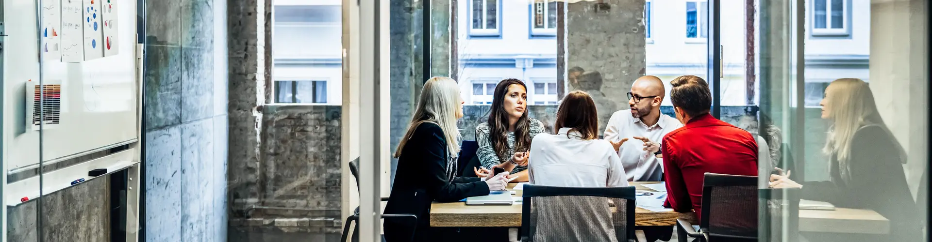 A group of people are sitting at a table during a business meeting in a bright, modern office with large windows and charts on the wall
