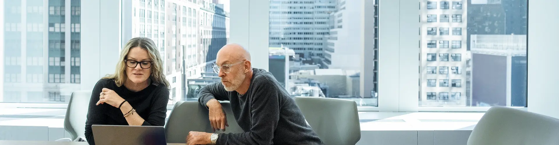 A man and woman are in a conference room with large windows, sitting at the table, having a conversation while looking at a laptop computer.