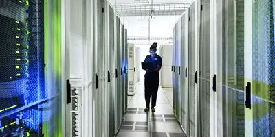 woman is walking down a hallway in a server room looking at her laptop