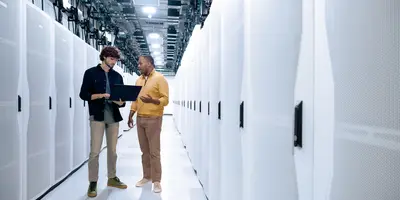 Two men standing in a server room looking at a laptop.