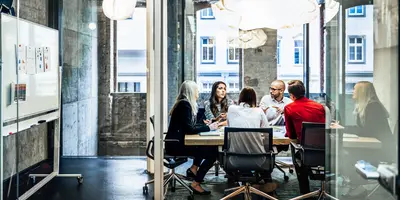 A group of people are sitting at a table during a business meeting in a bright, modern office with large windows and charts on the wall