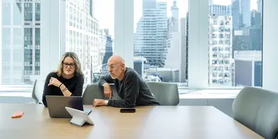 A man and woman are in a conference room with large windows, sitting at the table, having a conversation while looking at a laptop computer.
