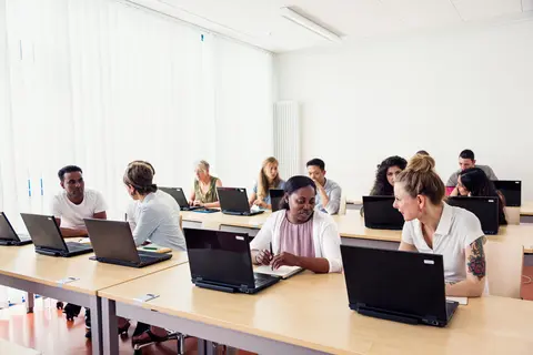 group of people are sitting at tables with laptops in a classroom
