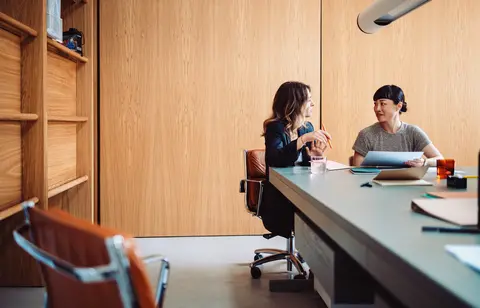 Two businesswomen engaged in a discussion at a modern office table. They are collaborating on a project with documents and laptops.