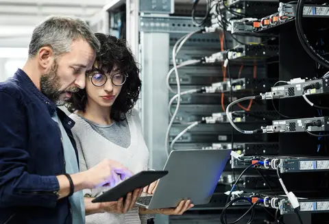 Male engineer showing a digital tablet to female coworker in a server room.