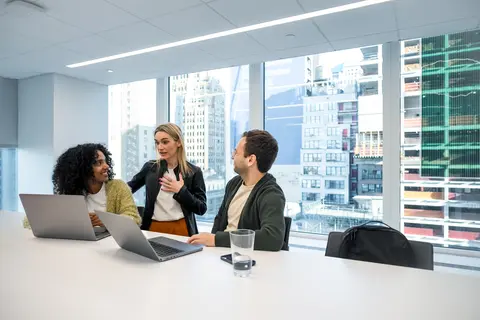 A group of people are sitting at a table with laptops having a conversation.
